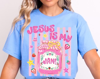 May include: A light blue t-shirt featuring a graphic print of a pink jam jar with a checkered lid. The text reads "JESUS IS MY JAM!" in pink letters. The design includes small illustrations of red strawberries, yellow stars, and pink flowers against a background of vertical pink stripes.
