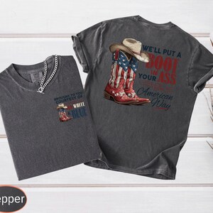 May include: Two dark gray t-shirts with a Western theme. One shirt features a graphic of cowboy boots with an American flag design and the text "We'll put a boot in your ass American Way." The other shirt has a smaller boot graphic and the text "Brought to you courtesy of White & Blue."