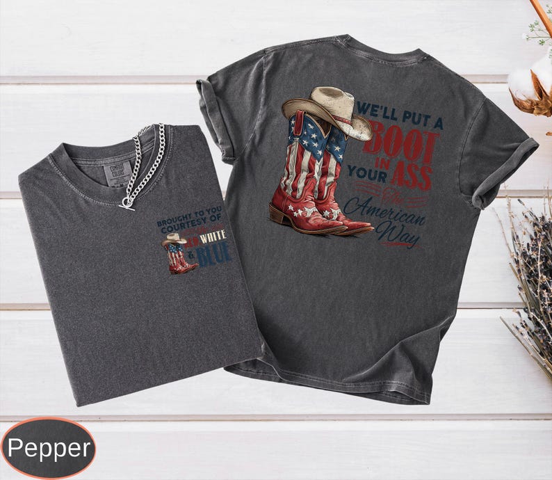 May include: Two dark gray t-shirts with a Western theme. One shirt features a graphic of cowboy boots with an American flag design and the text "We'll put a boot in your ass American Way." The other shirt has a smaller boot graphic and the text "Brought to you courtesy of White & Blue."