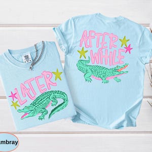 Puede incluir: Dos camisetas azul claro con dise&ntilde;os a juego. Una presenta la palabra "LATER" en rosa con un gr&aacute;fico de un caim&aacute;n verde. La otra tiene "AFTER WHILE" en la espalda, tambi&eacute;n con un gr&aacute;fico de caim&aacute;n.