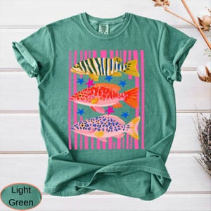 May include: Light green t-shirt featuring a colorful graphic of three fish. The fish are set against a pink striped background with blue stars and pink star accents. The shirt has a crew neck and short sleeves.