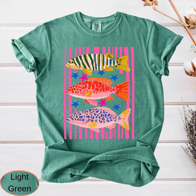 May include: Light green t-shirt featuring a colorful graphic of three fish. The fish are set against a pink striped background with blue stars and pink star accents. The shirt has a crew neck and short sleeves.