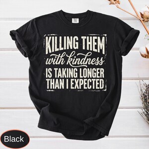 Op de afbeelding: Een zwart t-shirt met korte mouwen en witte tekst in een verweerde stijl met de tekst: KILLING THEM with kindness IS TAKING LONGER THAN I EXPECTED. Het shirt heeft een ronde hals en een standaard pasvorm, met de afbeelding gecentreerd op de borst.
