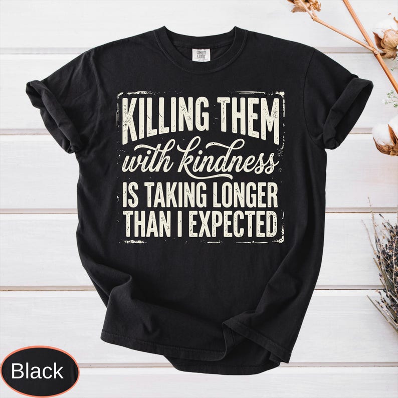 Op de afbeelding: Een zwart t-shirt met korte mouwen en witte tekst in een verweerde stijl met de tekst: KILLING THEM with kindness IS TAKING LONGER THAN I EXPECTED. Het shirt heeft een ronde hals en een standaard pasvorm, met de afbeelding gecentreerd op de borst.