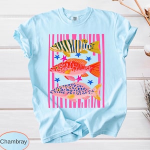 A garment-dyed shirt graphic featuring three vibrant, patterned fish in yellow, red, and pink over bold black and hot pink vertical stripes with scattered playful stars.