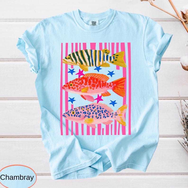 A garment-dyed shirt graphic featuring three vibrant, patterned fish in yellow, red, and pink over bold black and hot pink vertical stripes with scattered playful stars.