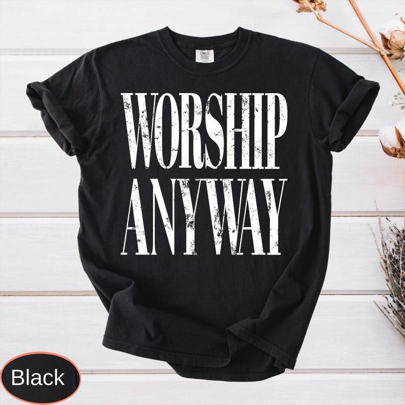 May include: Black t-shirt with the words "WORSHIP ANYWAY" printed in large, distressed white letters. The shirt is made of a soft material and has a classic crew neck. The shirt is displayed on a white wooden surface.