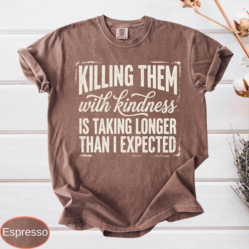 Op de afbeelding: Een bruin t-shirt met korte mouwen en de cr&egrave;mekleurige tekst "KILLING THEM with kindness IS TAKING LONGER THAN I EXPECTED" in een mix van blok- en scriptfonts op de voorzijde. Het shirt heeft een ronde hals en een relaxte pasvorm.