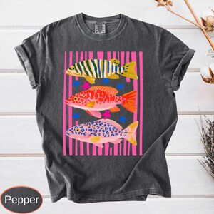 May include: Dark gray t-shirt featuring a colorful fish design. The graphic includes three stylized fish in yellow, orange, and pink, set against a pink striped background with blue stars. The shirt has rolled-up sleeves.