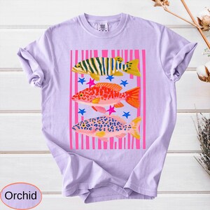 May include: A light purple t-shirt featuring a colorful graphic of three fish. The fish are set against a pink striped background with blue and pink star accents. The word "Orchid" is in a light purple oval at the bottom.