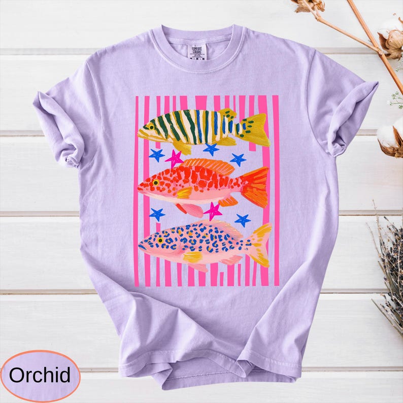 May include: A light purple t-shirt featuring a colorful graphic of three fish. The fish are set against a pink striped background with blue and pink star accents. The word "Orchid" is in a light purple oval at the bottom.