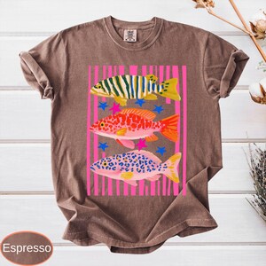 May include: Espresso-colored t-shirt featuring a colorful graphic of three fish. The fish are in shades of yellow, orange, pink, and blue, set against a pink striped background with blue stars. The shirt has short sleeves and a crew neck.