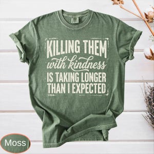 Op de afbeelding: Een mosgroen t-shirt met korte mouwen en de witte tekst "KILLING THEM with kindness IS TAKING LONGER THAN I EXPECTED" in een combinatie van blokletters en cursief schrift op de borst. Het kledingstuk heeft een ronde hals en een standaard pasvorm.