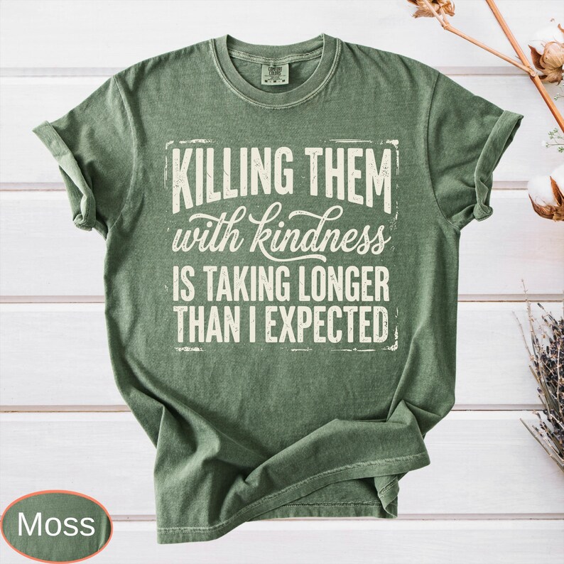 Op de afbeelding: Een mosgroen t-shirt met korte mouwen en de witte tekst "KILLING THEM with kindness IS TAKING LONGER THAN I EXPECTED" in een combinatie van blokletters en cursief schrift op de borst. Het kledingstuk heeft een ronde hals en een standaard pasvorm.