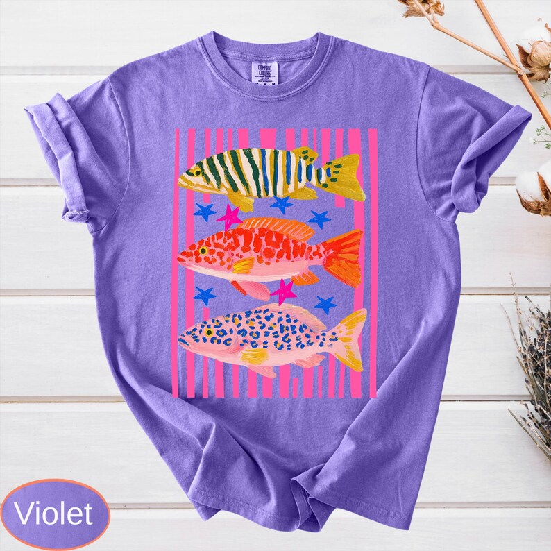 May include: Violet-colored t-shirt featuring a colorful fish design. The graphic includes three distinct fish illustrations with varying patterns and colors, set against a pink striped background with blue and pink star accents. The shirt has a classic crew neck.