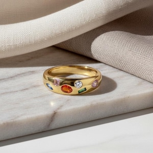 May include: A gold band ring adorned with various coloured gemstones. The ring showcases an oval orange stone, a round clear stone, and other stones in pink, blue, and green. It rests on a white marble surface.
