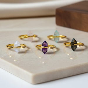 Könnte beinhalten: Eine Sammlung goldfarbener Ringe, jeder mit einem unterschiedlich farbigen Kristall. Die Kristalle sind in den Farbtönen Weiß, Rosa, Lila, Blau und Grau gehalten. Die Ringe werden auf einer hellen, quadratischen Oberfläche präsentiert.