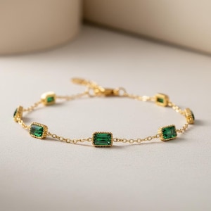 Puede incluir: Una delicada pulsera dorada con piedras preciosas rectangulares de color verde esmeralda. La pulsera tiene una fina cadena y es ajustable. Las piedras preciosas están engastadas en un engaste de bisel dorado. La pulsera se muestra sobre un fondo neutro.