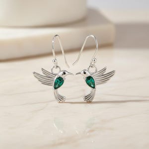 May include: A pair of silver hummingbird earrings with green gemstone accents. The earrings feature detailed wings and a delicate hook design. The gemstones are emerald-cut and add a pop of color to the jewelry.