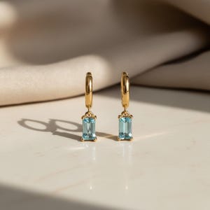 May include: A pair of gold hoop earrings with rectangular, light blue gemstones. The earrings are displayed on a white surface with a soft, neutral background. The gemstones are a light, clear blue color.