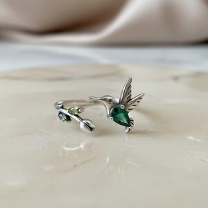 May include: A silver open ring featuring a hummingbird design with a pear-shaped green gemstone body. The opposite end of the band depicts a small silver branch with two marquise-cut olive green stones and a small silver flower bud detail.
