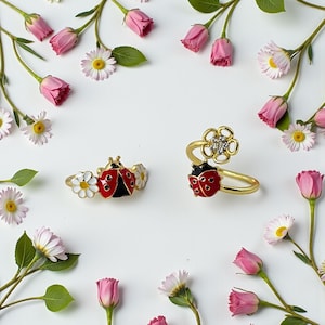 May include: Two gold-toned rings featuring ladybug motifs. One ring displays a red and black enamel ladybug flanked by white daisy flowers. The second ring features a red and black enamel ladybug paired with a gold flower set with clear stones.