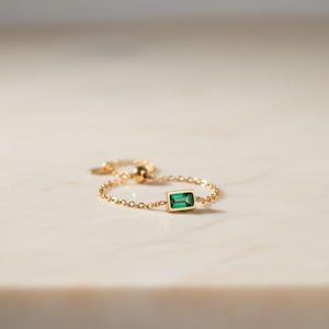 May include: A delicate gold chain ring featuring a rectangular emerald-cut green gemstone. The ring has a fine chain band and an adjustable clasp. The ring is set against a neutral background.