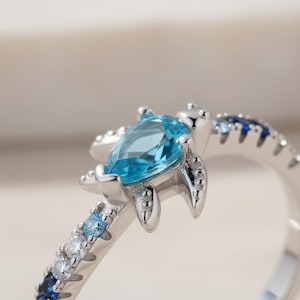 Peut inclure: Une bague en argent avec un motif de tortue orné d'une pierre précieuse bleu clair en forme de poire. L'anneau est décoré d'un dégradé de petites pierres rondes, passant du bleu foncé au bleu clair et au blanc transparent sur la tige.