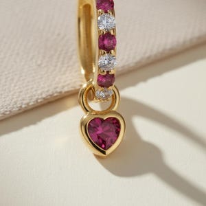 Può includere: Un orecchino a cerchio dorato con un ciondolo a forma di cuore. Il cerchio è adornato da pietre preziose rosa e bianche alternate. Il ciondolo a forma di cuore è di un rosa intenso ed è incastonato in una montatura dorata. L'orecchino è esposto su una superficie color crema.