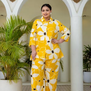 Può includere: Un completo a due pezzi composto da una camicia abbottonata con maniche a tre quarti e pantaloni a gamba larga. Il capo presenta uno sfondo giallo vivace con un motivo floreale bianco su larga scala e centri circolari marrone scuro.