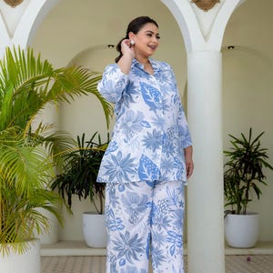 May include: A two-piece pajama set with a white base and a blue floral design. The set comprises a button-down shirt and wide-leg trousers. The model is wearing the set outdoors, with plants and architectural features in the background.