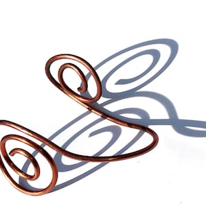 May include: A copper arm cuff featuring a spiral design. The cuff curves around the arm with two prominent spiral accents. The piece is made of a single piece of metal and casts a shadow on a white surface.