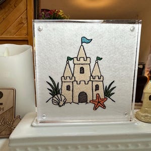 May include: A square acrylic frame containing a white fabric insert with an embroidered sandcastle design. The embroidery features a beige castle with blue and green flags, a brown doorway, a tan seashell, a red starfish, and green grass.