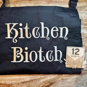 May include: Black apron with the words "Kitchen Bitch" embroidered in a gold-colored script. A small wooden emblem with the text "12 North Embroidery" is attached to the apron.