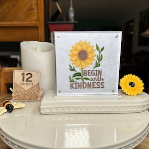 May include: A decorative display featuring a sunflower embroidery with the text "Begin with Kindness." A small wooden sign with "12 North Embroidery" is also present, along with a bee figurine and a yellow sunflower.