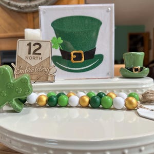 May include: A St. Patrick's Day themed display featuring a green shamrock, a beaded garland with green, white, and gold beads, and a green leprechaun hat. A sign with the text "12 North Embroidery" is also present.