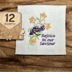 May include: A wooden sign with the number 12 and the word "NORTH" above "Embroidery" in script. Next to it is a white square with an embroidered cross, flowers, and the words "Risen" and "Rejoice in our Saviour" in purple.