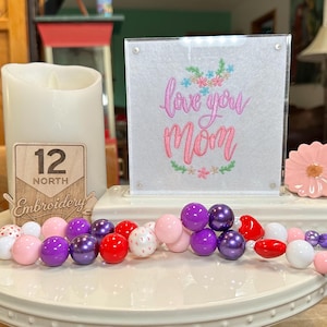 May include: A decorative display featuring a white candle, a sign that says "love you Mom", a pink flower, and a beaded garland with purple, pink, red, and white beads. The display is on a white ceramic tray.