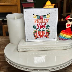 May include: A white tiered display stand featuring a square sign with the embroidered text "FEELIN' Hot" above two red chili peppers and colorful bunting. A white pillar candle sits on the left, and a small figurine in a red and rainbow dress sits on the right.