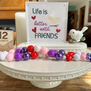 May include: A decorative beaded garland with purple, pink, red, and white beads, and a purple tassel. A sign with the text "Life is better with friends" and a small ceramic bird are on a white cake stand.