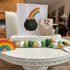 May include: A St. Patrick's Day themed display featuring a rainbow, a pot of gold, and a beaded garland. The display includes a framed embroidery piece, a wooden sign that says "12 North Embroidery", and a small pot of gold.