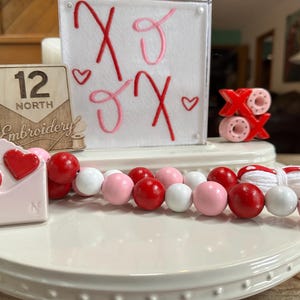 May include: A Valentine's Day-themed display featuring a beaded garland with red, pink, and white beads. The display includes a sign with "XO" and hearts, a wooden sign, and a decorative envelope with hearts.