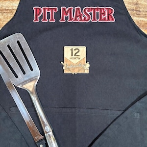 May include: Black apron with the words "PIT MASTER" in red and white lettering. A metal spatula and knife are in the pocket. A wooden emblem with the text "12 NORTH Embroidery" is also on the apron.