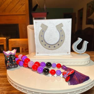 May include: A decorative display featuring a horseshoe theme. It includes a framed horseshoe design, a small horseshoe figurine, and a beaded garland with red, purple, and black beads. A small boot-shaped vase is also present.