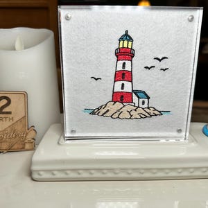 May include: A square acrylic frame containing a white fabric panel with an embroidered red and white lighthouse on a rocky base, featuring black birds in the sky. A small lighthouse figurine and a wooden sign reading 12 NORTH Embroidery are present.