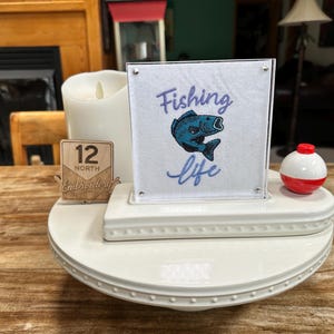 May include: A white square sign featuring the embroidered text Fishing life and a blue fish graphic, displayed in a clear frame on a white tiered stand. Next to it is a small wooden sign labeled 12 North Embroidery, a white pillar candle, and a red and white fishing bobber.