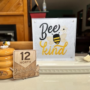 May include: A decorative display featuring a ceramic honey pot with a bee figurine, a wooden sign with "12 NORTH Embroidery" text, and a sign that says "Bee kind" with a bee graphic. The display is on a white tray.