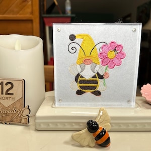 May include: A decorative display featuring a gnome dressed as a bee, holding a pink flower. The gnome is embroidered on a white background, set in a clear acrylic frame. A wooden sign reads "12 North Embroidery". A ceramic bee figurine is in the foreground.