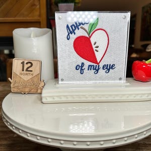 May include: A square white fabric panel featuring an embroidered red apple half and the blue text "Apple of my eye" displayed on a white tiered stand. The display includes a small wooden sign with "12 NORTH Embroidery" and a red ceramic apple.