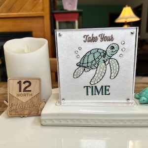 May include: A square white sign featuring an embroidered light blue sea turtle and the text "Take Your TIME" in dark blue. The sign is displayed on a white base next to a white pillar candle, a small wooden sign, and a small turquoise turtle.